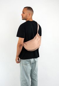 Borsa a tracolla rosa chiaro realizzata in tessuto morbido, forma curvata con una fettuccia sottile, dotata di chiusura con zip e un branding discreto.