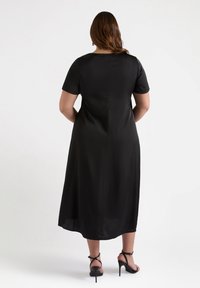 Vestido negro de longitud media con mangas cortas, textura suave y brillo sutil. Presenta una costura trasera recta. Se lleva con sandalias de tacón negras.