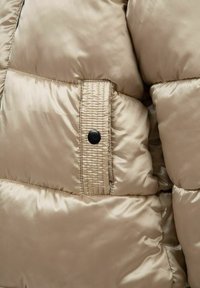 Beige pufferjakke med glansfuld stof, quiltet tekstur og en sort trykknap. Har horisontale sømme og en sidelomme.