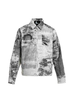 Hombre con rastas que lleva una chaqueta y pantalones con un patrón abstracto gris y blanco, camisa negra y zapatos marrones, de pie sobre un fondo blanco.