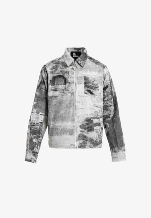 Hombre con rastas que lleva una chaqueta y pantalones con un patrón abstracto gris y blanco, camisa negra y zapatos marrones, de pie sobre un fondo blanco.