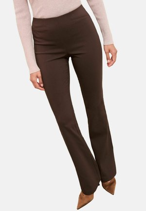 Frau trägt hoch taillierte, dunkelbraune Schlaghosen, kombiniert mit einem hellrosa Langarmtop und braunen spitz zulaufenden Absätzen.