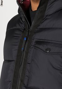 Černá péřová bunda s vysokým límcem, zapínáním na zip s modrým akcentem a vnější kapsou na hrudi s černým knoflíkem.