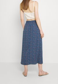 Maxihame sinisessä kankaassa, jossa on kukkakuosi; siinä on joustava vyötärönauhka ja väljä istuvuus. Yhdistetty beigen sävyiseen lyhytpaitaiseen toppiin ja sandaaleihin.