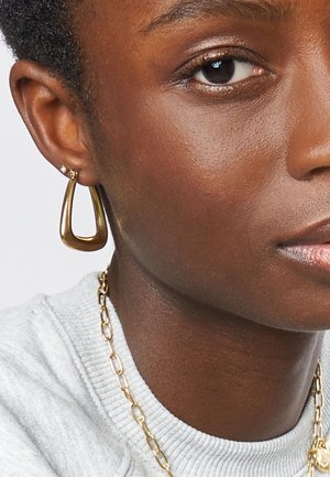 Goldene Creolen in offener Tränenform, mit glatter Oberfläche und polierter Verarbeitung, getragen von einer Person mit kurzen Haaren und einem grauen Pullover.
