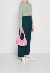 Kvinde iført en langærmet mintgrøn crop top, mørke tealfarvede vide bukser, hvide sneakers, mens hun holder en pink rynket taske ved siden af.