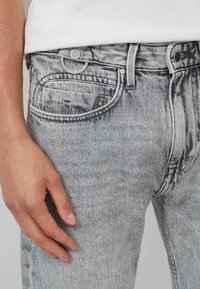 Szare dżinsy denimowe z klasycznym fasonem pięciu kieszeni, widocznym przeszyciem i zapięciem na guzik. Ręka spoczywa na udzie.