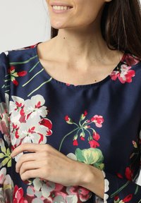 Blusa de seda con fondo azul marino y patrones florales coloridos en rojo, blanco y verde. Cuello redondo y mangas de tres cuartos.