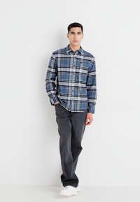 Camisa de cuadros azul de botones con detalles en blanco, azul marino y negro, combinada con jeans oscuros y zapatillas blancas. Mangas cortas y ajuste relajado.