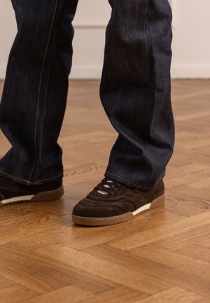 Persona che indossa jeans blu scuro e sneaker in suede nero, in piedi su un pavimento in parquet di legno.