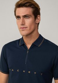 Polo de marina con mangas cortas, cuello con cremallera y ribete naranja en contraste. Logo "HACKETT" impreso en un color claro en la parte delantera.