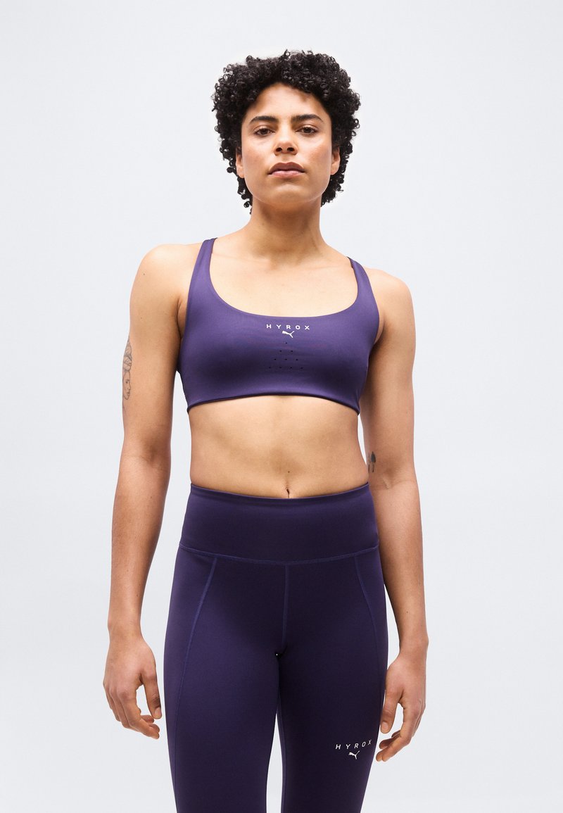 Γυναίκα με κοντά σγουρά μαλλιά, φορώντας μοβ αθλητικό σουτιέν και ψηλόμεσο κολάν, στέκεται μπροστά σε απλό φωτεινό φόντο.