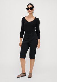Čierne priliehavé rebrované tričko s dlhými rukávmi a čipkovým lemom, čierne strihané šortky do kolien a zdobené čierne sieťované slip-on topánky.