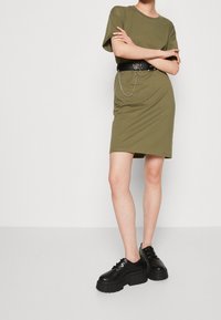 Oliwkowa sukienka T-shirt z krótkimi rękawami, dopasowana sylwetka, przewiązana w talii czarnym paskiem z łańcuszkiem, zestawiona z masywnymi czarnymi butami.