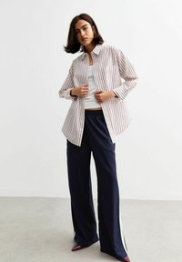 Gestreepte button-up shirt in wit en rood met lange mouwen en een borstzak, gecombineerd met marineblauwe wijde broeken en puntige bordeauxrode schoenen.