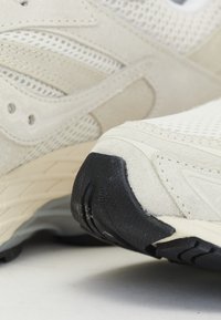 Scarpe sportive beige chiaro con tomaia in mesh e dettagli in suede. La suola è nera con un motivo testurizzato per garantire grip. Presenti dettagli di design sottili.