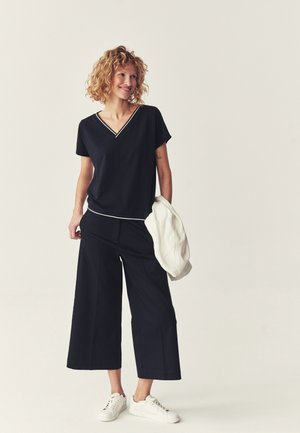 Vrouw met krullend haar, draagt een zwart V-hals topje, brede broekspijpen, witte sneakers, houdt een wit jasje vast, lacht en kijkt opzij.