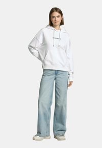 Felpa bianca con tasca a marsupio, caratterizzata da un logo nero, abbinata a jeans azzurri a gamba larga e sneakers neutre.