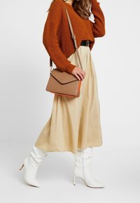 Borsa crossbody tan con un accento arancione, texture liscia e chiusura a busta. Indossata con una gonna crema e un maglione a maglia ruggine, completata da stivaletti bianchi.