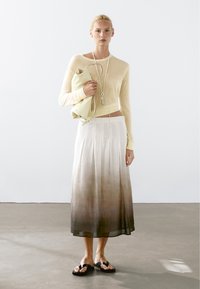 Beige gebreide trui, witte rok met een bruine gradient, en zwarte slippers. Houdt een lichtgekleurde tas vast, die een gladde textuur en een eenvoudig ontwerp heeft.