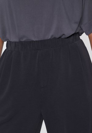 Záběr na osobu v černých elastických kalhotách s vysokým pasem a zasunutou tmavě šedou košilí, zaměřený na pas a horní část boků.