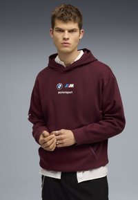 Sudadera burdeos con un logo, que presenta una capucha con cordón y puños acanalados. Superpuesta sobre una camisa blanca, con detalles en azul y negro.
