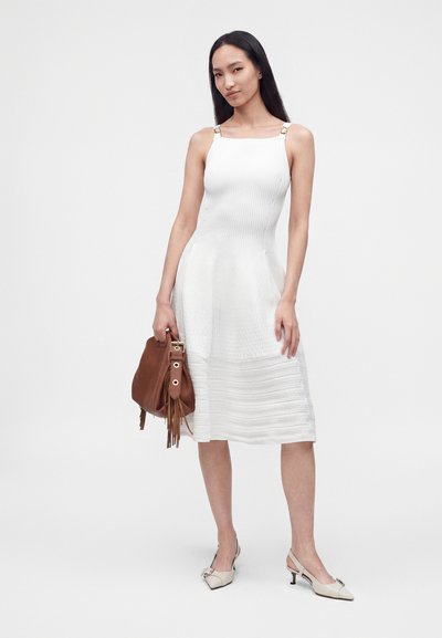 Femme portant une robe blanche sans manches et des talons beiges, tenant un sac à main marron avec des pompons, debout devant un fond uni clair.