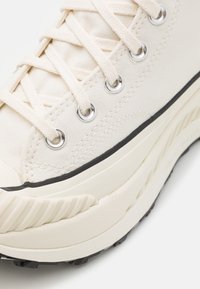 Beige canvas sneakers med platta snören, silverfärgade ögla, och en tjock, texturerad gummisul med en svart accentrand och spårigt mönster.