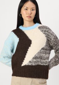 Sweter robiony na drutach z niebieskim kołnierzem, charakteryzujący się wyrazistymi zygzakowymi wzorami w kolorach brązowym, kremowym i szarym, wykonany z teksturowanego materiału wełnianego.