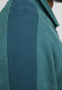 Strikket pullover med et tekstureret design. Har mørkegrønne og teal farveblokke med et fiskebensmønster på ærmerne.