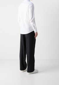 Persoon gezien vanaf de achterkant, gekleed in een wit langmouwshirt, zwarte wijde broeken en witte sneakers, staande tegen een effen achtergrond.