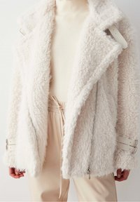 Chaqueta blanca y esponjosa con una superficie texturizada, detalles de cremallera plateada y tirantes ajustables en las muñecas. Diseño en capas con solapas anchas.