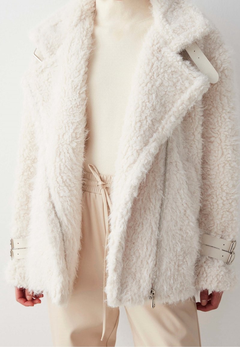 Chaqueta blanca y esponjosa con una superficie texturizada, detalles de cremallera plateada y tirantes ajustables en las muñecas. Diseño en capas con solapas anchas.
