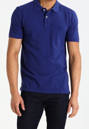 Uomo che indossa una polo blu aderente a maniche corte con colletto e pantaloni scuri, in piedi di fronte a uno sfondo bianco.