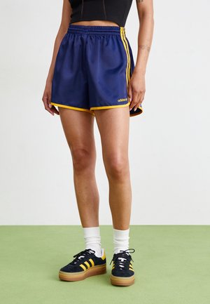 Personne portant un short Adidas bleu marine avec des bandes jaunes, des baskets noires et jaunes, des chaussettes blanches et un top court noir, debout sur un sol vert.