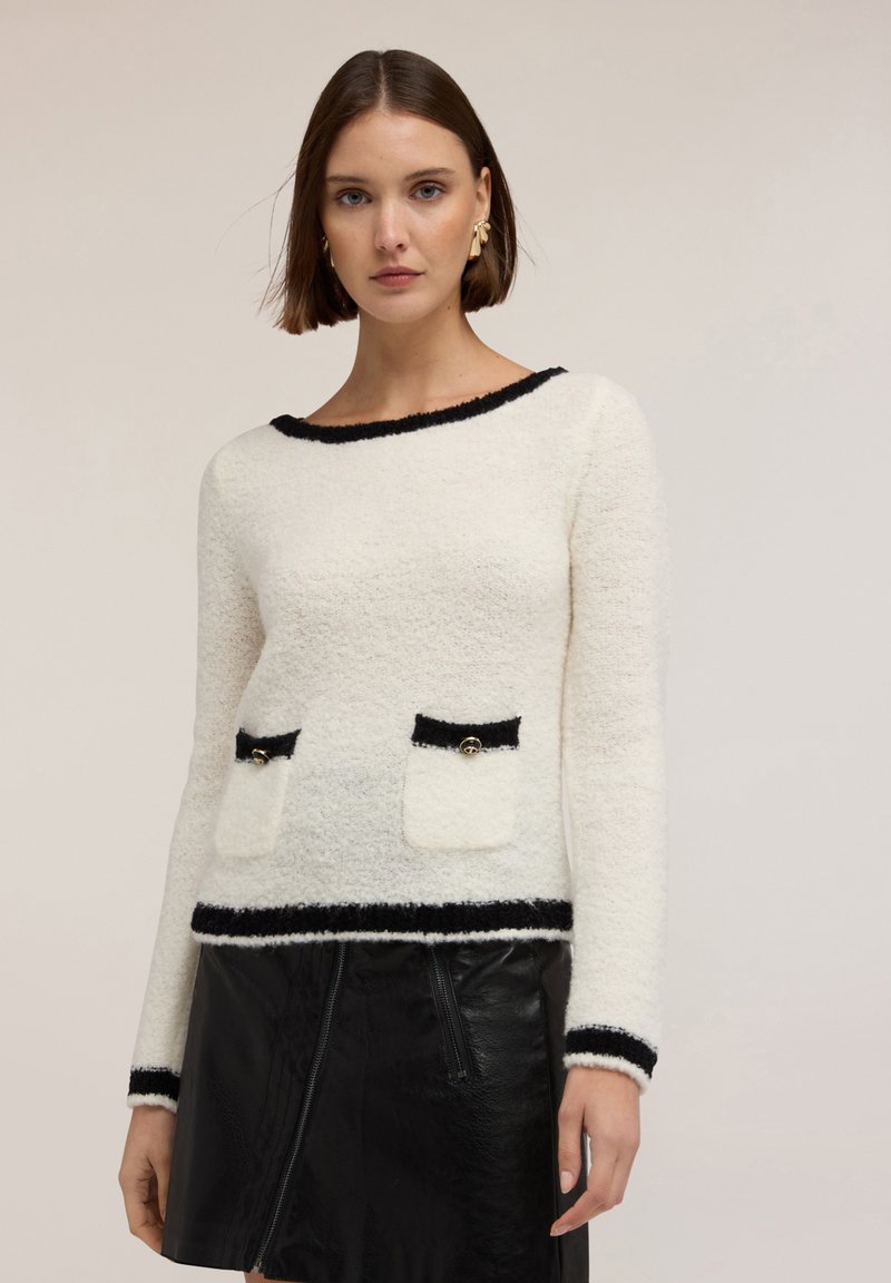 Donna che indossa un maglione bianco a maniche lunghe con texture e bordi neri, abbinato a una minigonna in pelle nera su uno sfondo neutro.