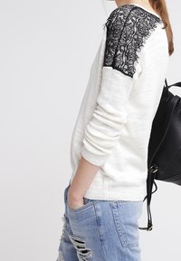 Femme portant un pull blanc avec un détail en dentelle noire sur l'épaule, un jean bleu déchiré et un sac à dos en cuir noir.