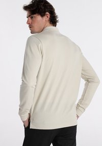 Langärmliges, kragenloses Poloshirt in hellem Beige mit strukturiertem Stoff. Das Shirt verfügt über seitliche Schlitze und wird mit dunklen Hosen kombiniert.