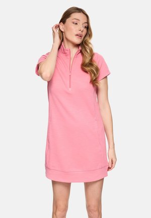 Mujer con cabello largo y ondulado, vestida con un vestido rosa de manga corta con cremallera frontal, posando frente a un fondo liso y claro.