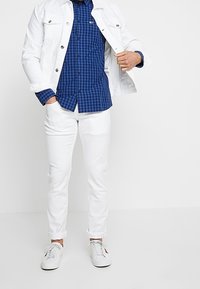 Biała kurtka dżinsowa, niebieska koszula w kratę, białe spodnie i białe trampki. Kurtka zapinana na guziki, mankiety podwinięte, a na koszuli znajduje się logo.