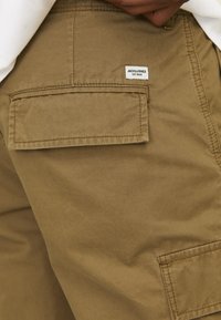 Shorts cargo marron avec une grande poche arrière et une étiquette de marque blanche portant l'inscription "JACK & JONES EST. 1990." Tissu texturé et résistant.