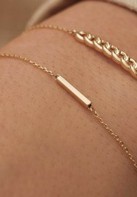 Bracelet en or avec un pendentif rectangulaire sur une chaîne fine, sur un fond de texture en peau et un bracelet à maillons épais à proximité.