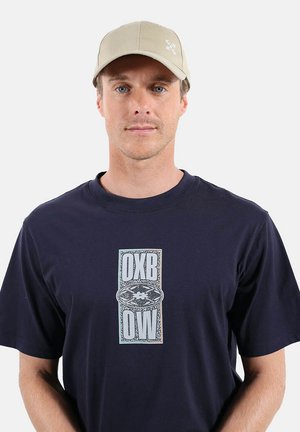 T-shirt bleu marine avec un graphisme centré affichant "OXB" au-dessus de "OW". Casquette beige avec logo. Tissu lisse, manches courtes, col rond.
