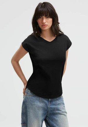 Woman with dark hair wearing a black short-sleeve V-neck shirt and blue jeans standing against a plain light background.