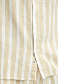 La camisa a rayas beige presenta rayas verticales blancas, está hecha de una tela ligera, cuenta con botones en la parte delantera y tiene una etiqueta en el lateral.