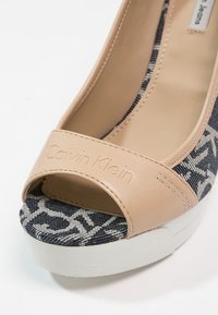 Sandaletta beige in pelle con tacco a cuneo, punta aperta, tomaia in tessuto stampato blu navy e bianco, e suola in gomma bianca. Logo Calvin Klein impresso.