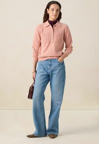 Pull-over tricoté rose clair avec col, patte de boutonnage et deux poches poitrine. Associé à un jean bleu évasé et des chaussures à motifs.