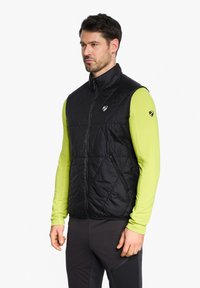 Black quilted vest with a high collar and front zipper, featuring a logo on the chest and side pockets, worn over a neon green long-sleeve shirt.