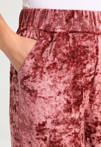Samt-Hosen in einem tiefen rosa Farbton verfügen über einen gerafften Bund und seitliche Taschen. Der Stoff hat eine weiche, strukturierte Oberfläche mit subtilen Mustern.