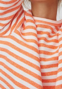 Oranssi-valkoraidallinen kangas, jossa pehmeä tekstuuri ja rento laskeutuvuus, vaakasuoria viivoja eri paksuuksilla.